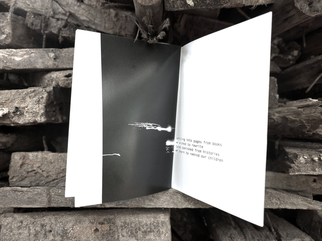 Open spread — dark page with white abstract mark and text reading "settling into pages from books we wished to rewrite, tales banished from histories we learn to remind our children", on charcoal wood