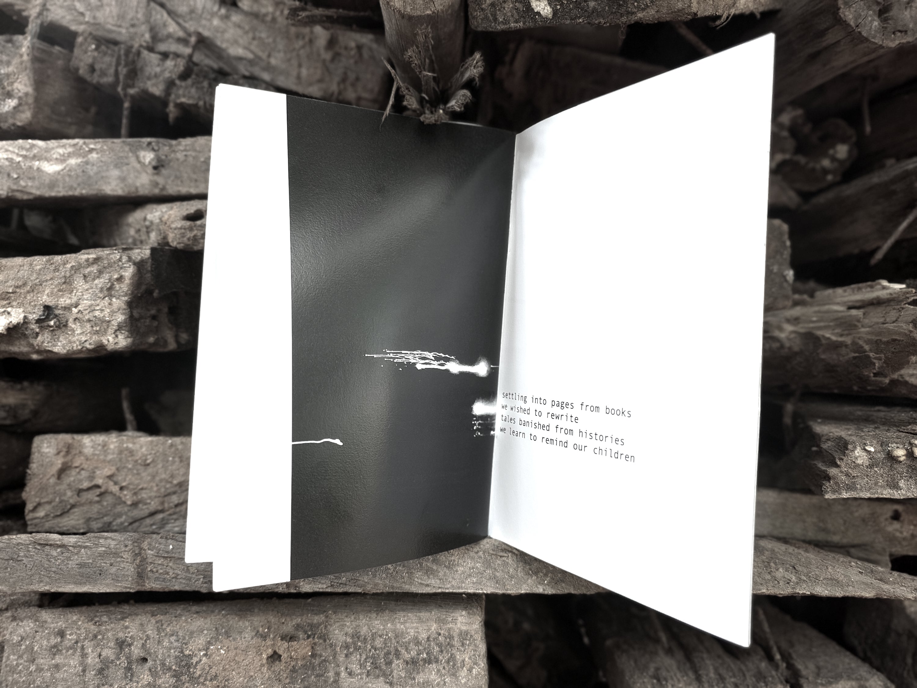 Open spread — dark page with white abstract mark and text reading "settling into pages from books we wished to rewrite, tales banished from histories we learn to remind our children", on charcoal wood