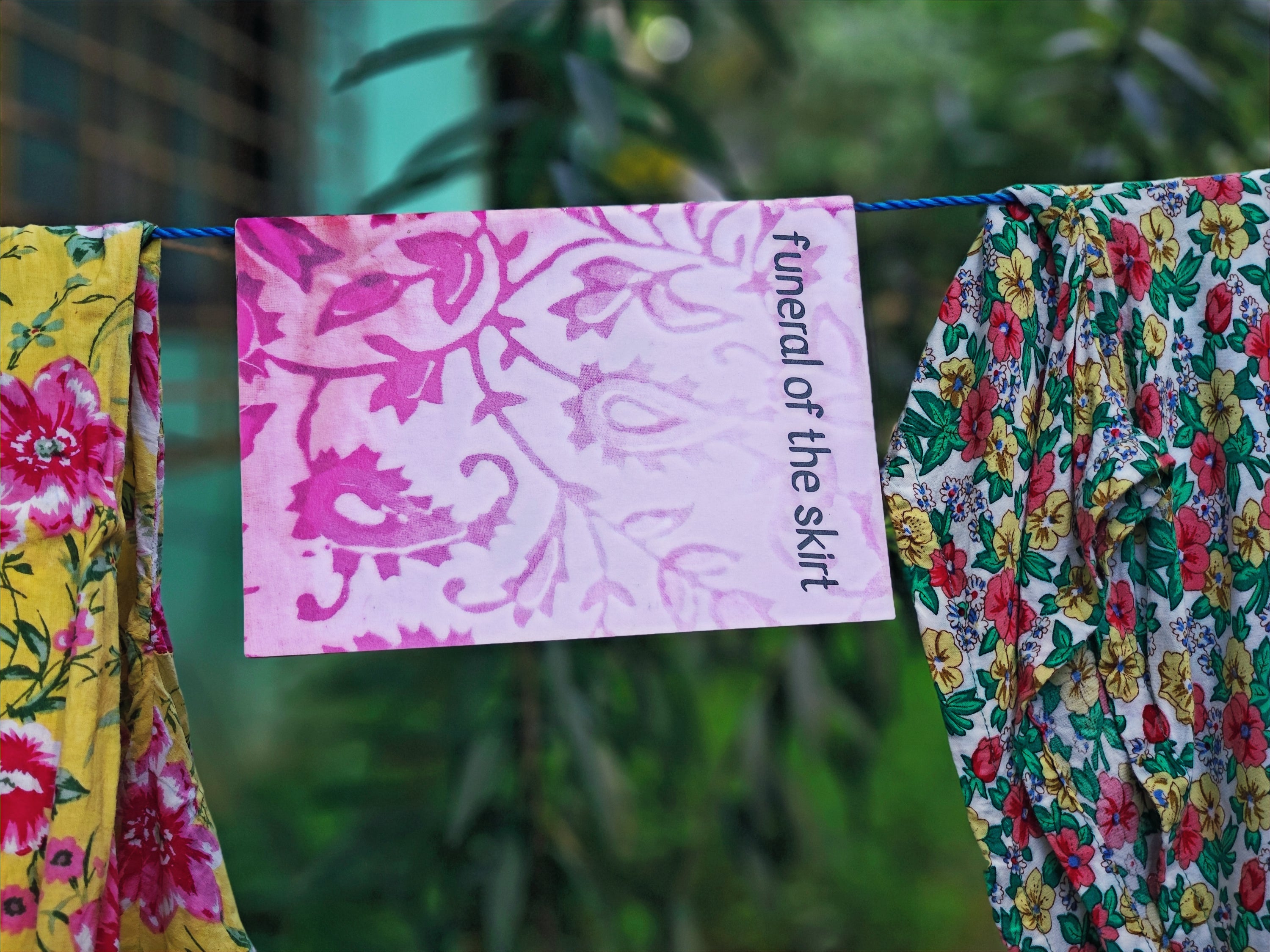 "Funeral of the Skirt" zine cover — pink paisley patterned cover with title text, hanging on a clothesline between yellow floral and white floral fabrics, green foliage behind