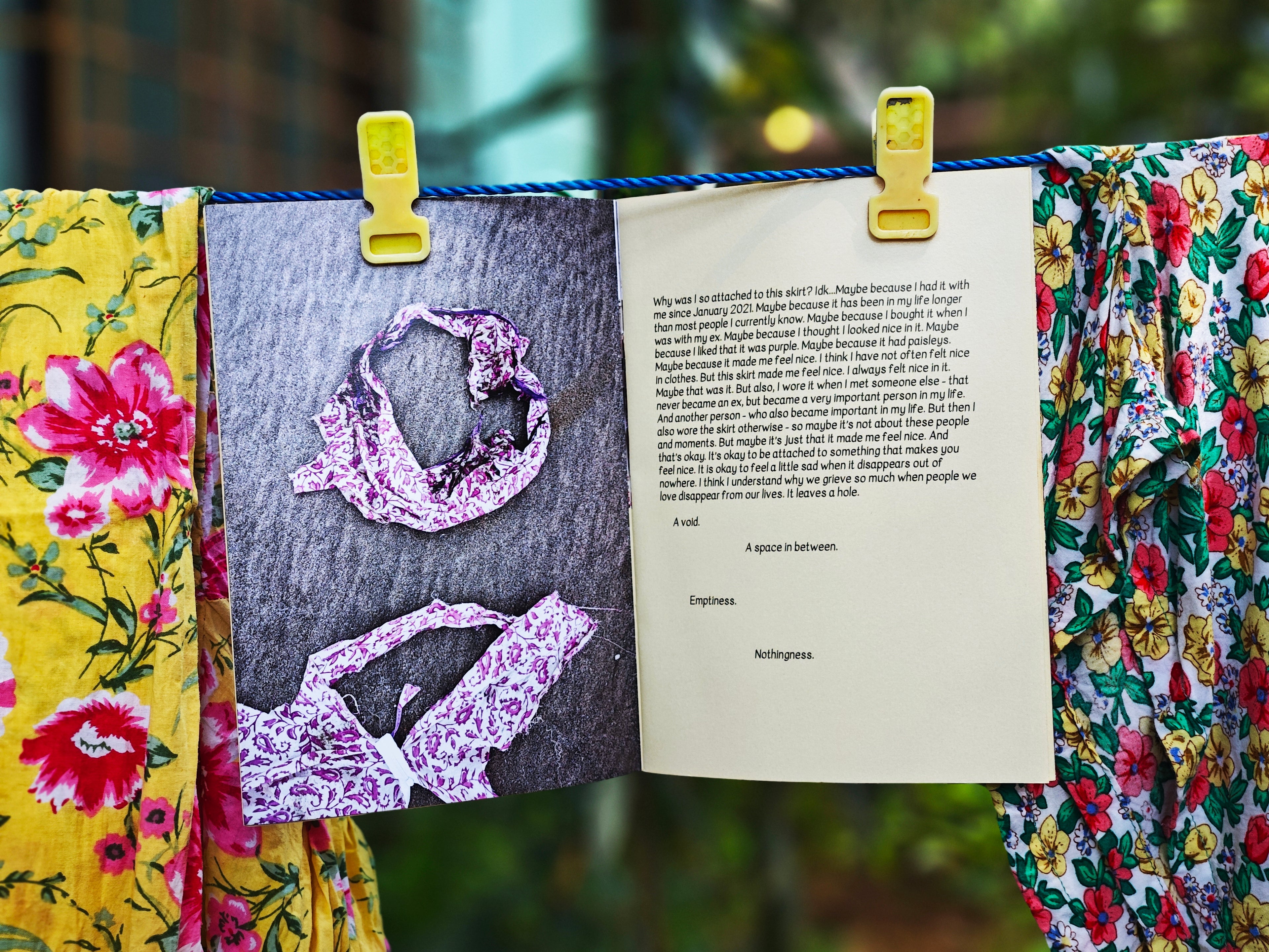 Open spread on clothesline — left page shows photographs of crumpled purple paisley skirt, right page with typed text about attachment to the skirt and words "a void, a space in between, emptiness, nothingness", hanging between floral fabrics