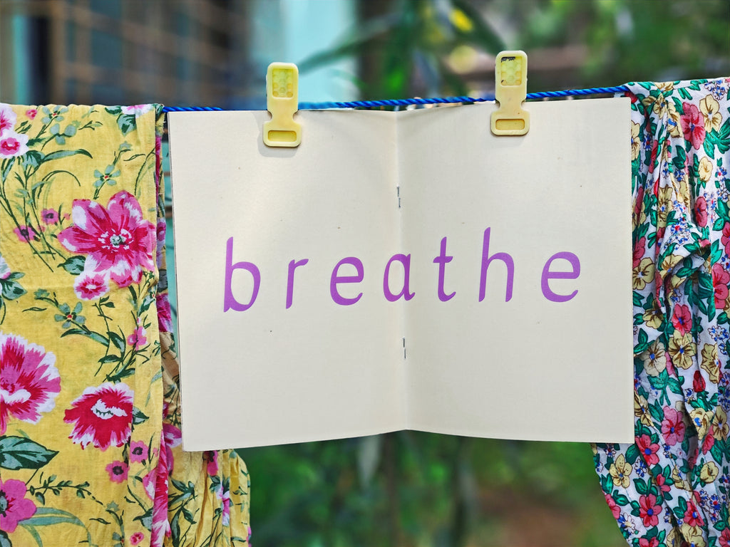 Open spread on clothesline — cream pages with large purple lowercase text reading "breathe", hanging between floral fabrics with green garden behind