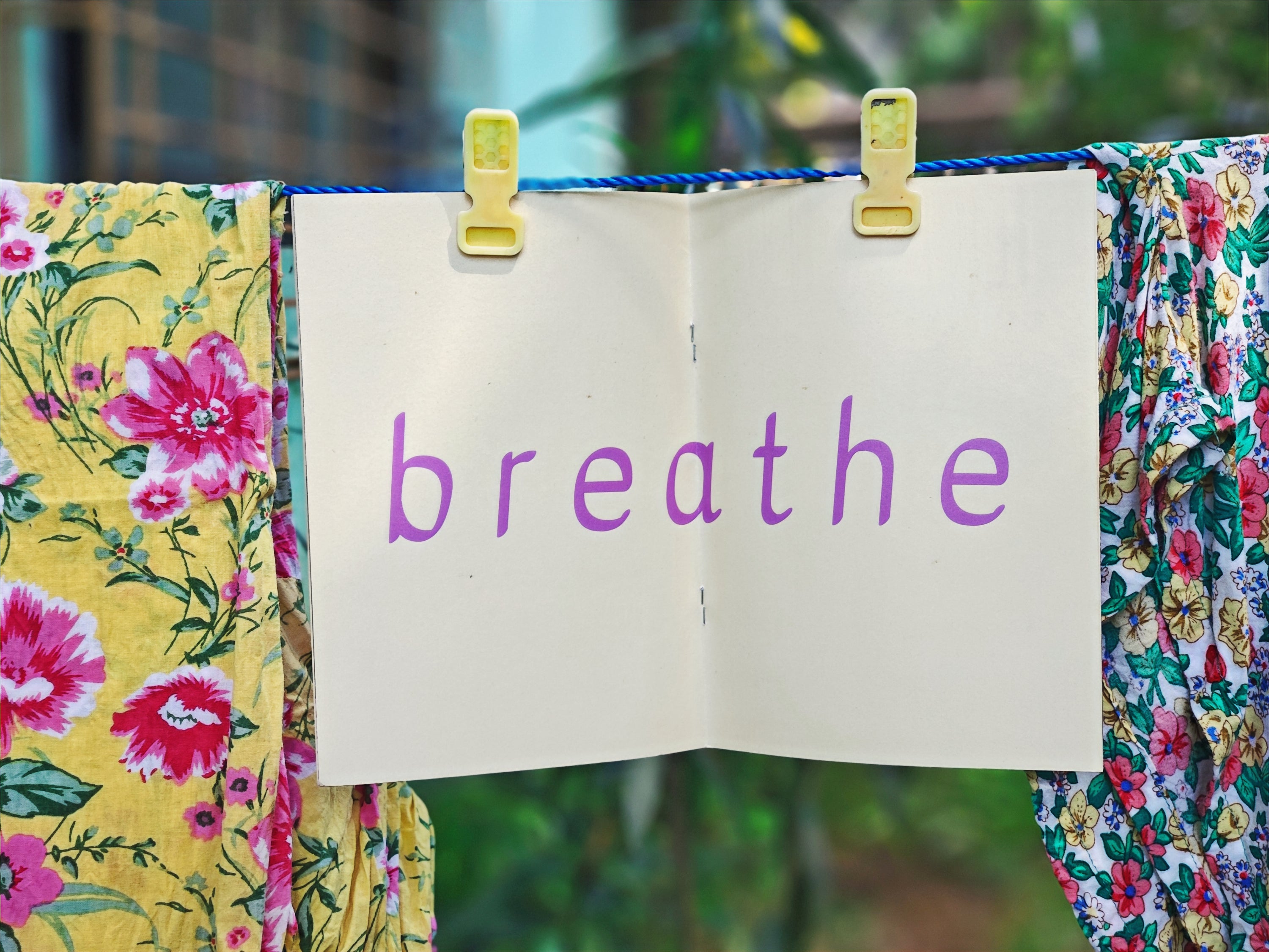 Open spread on clothesline — cream pages with large purple lowercase text reading "breathe", hanging between floral fabrics with green garden behind