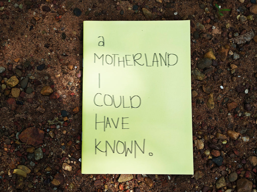 "A Motherland I Could Have Known" zine cover — handwritten title on pale green paper, resting on red Goan earth and pebbles