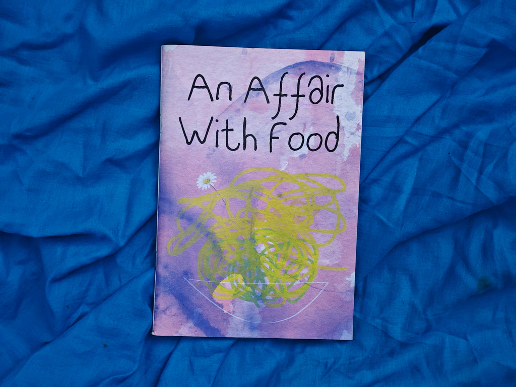 "An Affair With Food" zine cover — purple and yellow watercolour with abstract scribbles and a pressed daisy, resting on crumpled blue fabric