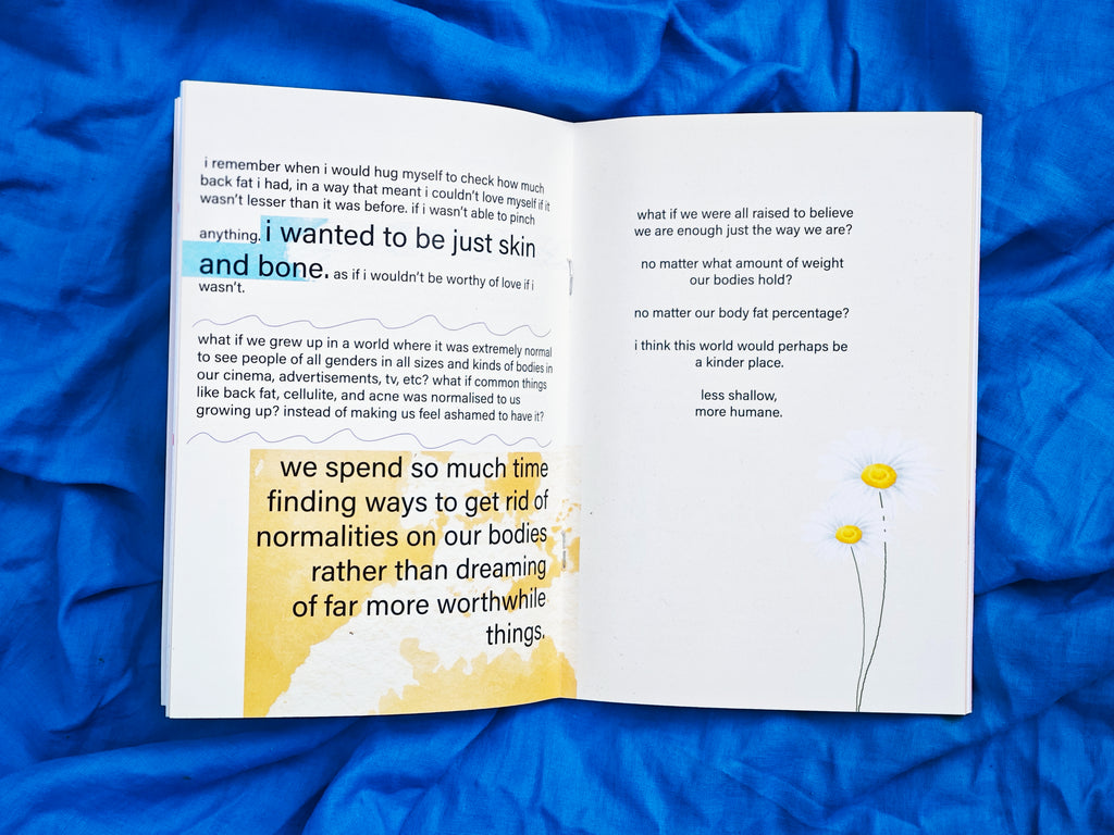 Open spread on blue fabric — left page with highlighted text "i wanted to be just skin and bone" and "we spend so much time finding ways to get rid of normalities on our bodies rather than dreaming of far more worthwhile things" — right page reads "what if we were all raised to believe we are enough just the way we are?" with daisy illustrations, "less shallow, more humane"