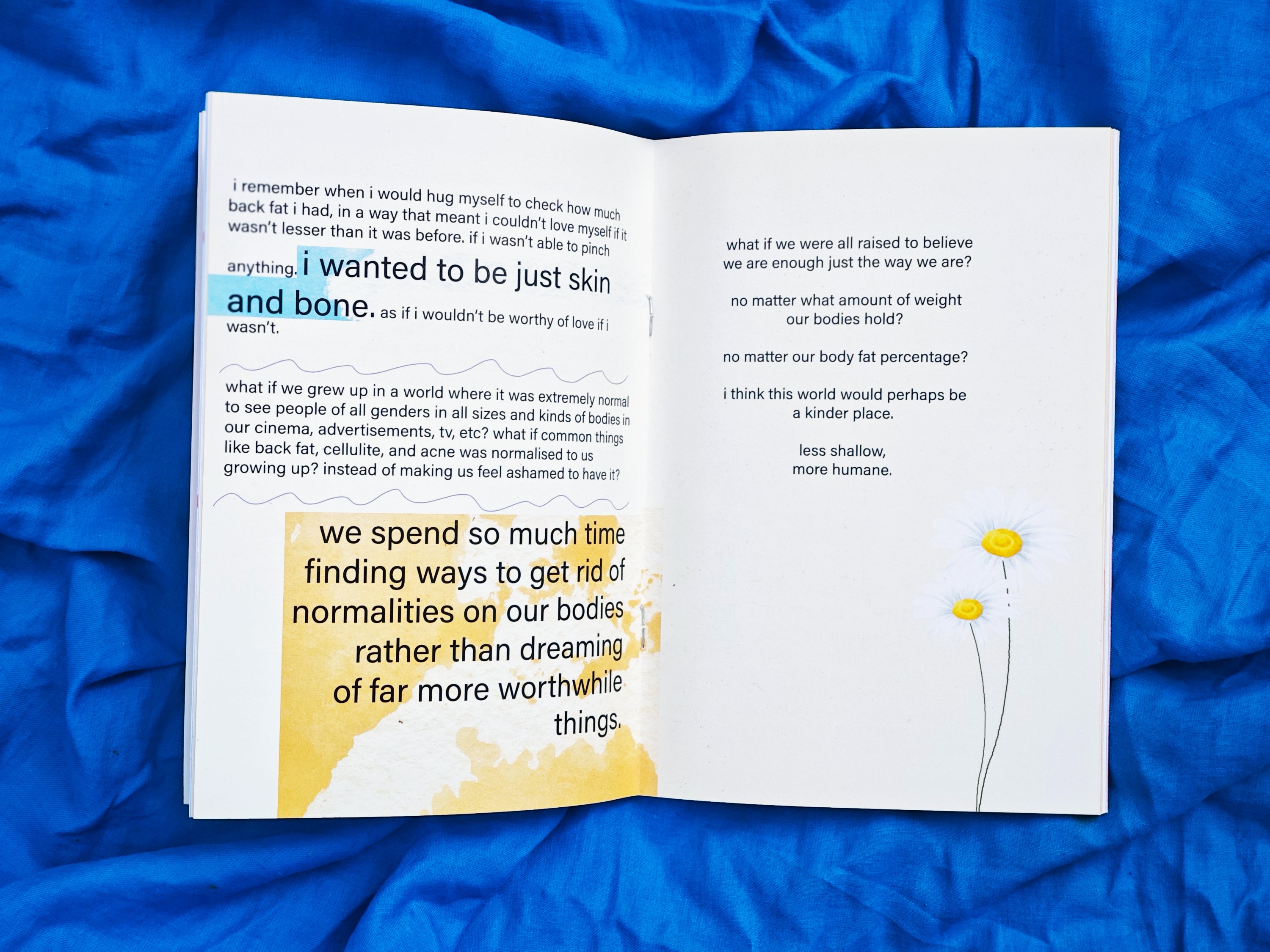 Open spread on blue fabric — left page with highlighted text "i wanted to be just skin and bone" and "we spend so much time finding ways to get rid of normalities on our bodies rather than dreaming of far more worthwhile things" — right page reads "what if we were all raised to believe we are enough just the way we are?" with daisy illustrations, "less shallow, more humane"