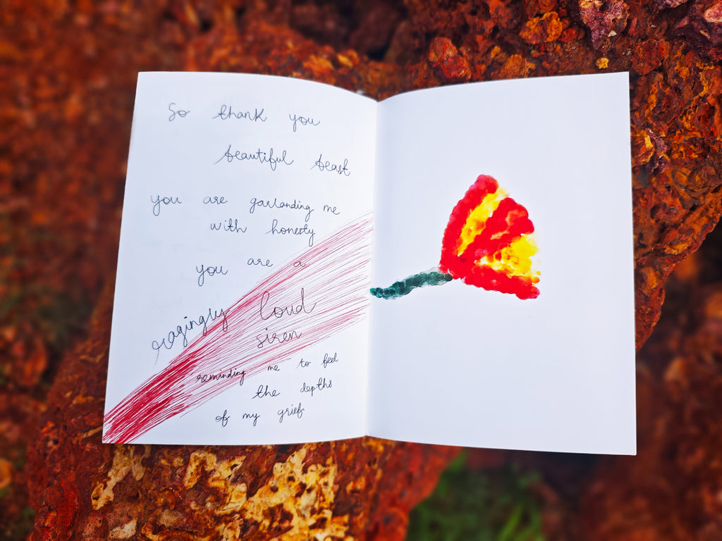 Open spread on laterite rock — left page with handwritten text "so thank you beautiful beast, you are garlanding me with honesty", right page with painted red and yellow flower