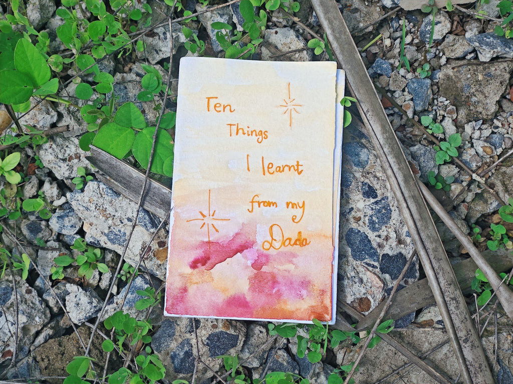 "Ten Things I Learnt From My Dada" — hand-painted watercolour cover in sunset pinks and oranges with handwritten title and small star drawings, resting on ground among green clover plants
