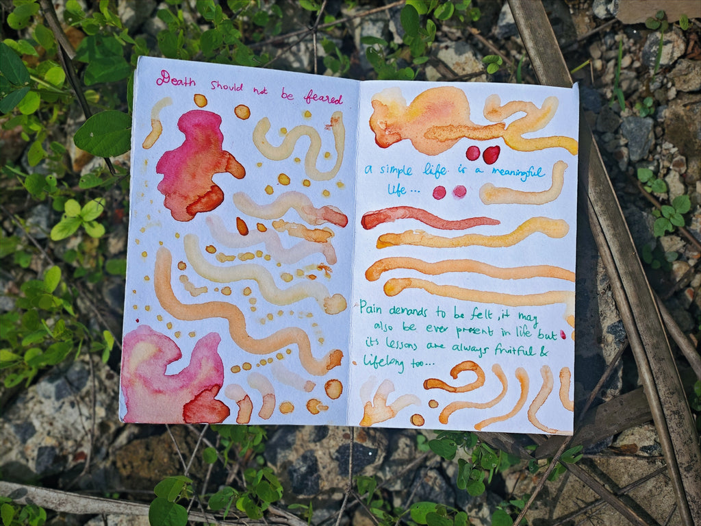 Open spread on ground among plants — handwritten text "Death should not be feared" and "a simple life is a meaningful life" with pink, orange and golden watercolour swirls and dots