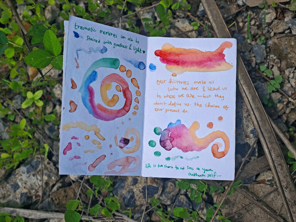 Open spread on ground among plants — handwritten text "traumatic memories can also be stained with goodness & light" and "our histories make us who we are" with colourful rainbow swirls and watercolour forms