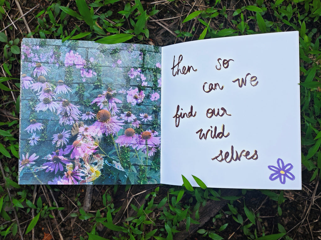 Open spread on grass — left page with photograph of pink and purple wildflowers growing through a wall, right page with handwritten text "then so can we find our wild selves" and small purple flower doodle