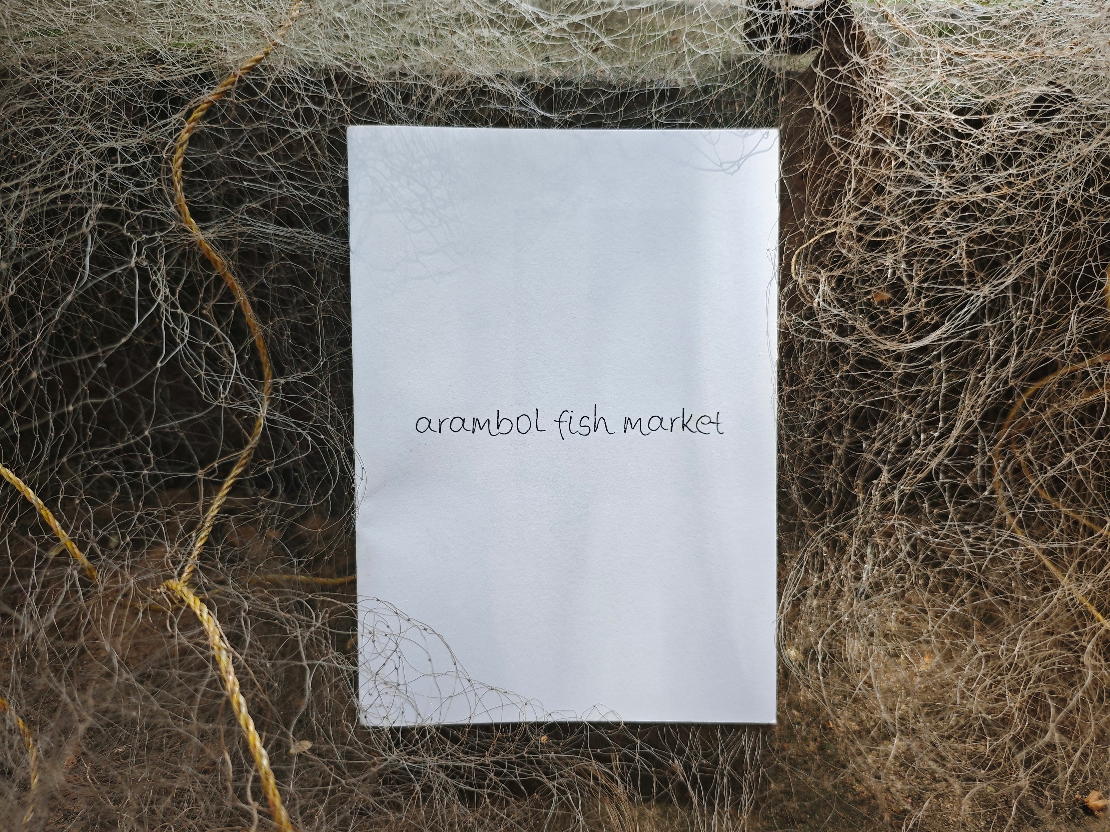 "Arambol Fish Market" zine cover — handwritten title on white paper, resting on tangled grey fishing nets with golden rope