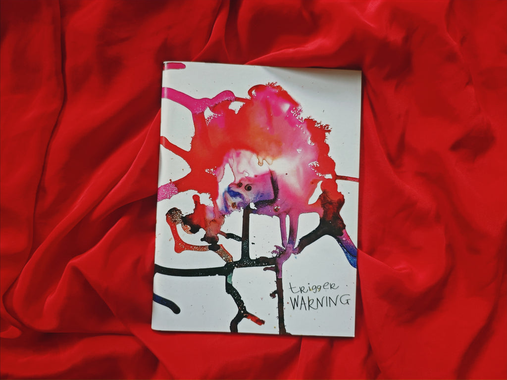 "Trigger Warning" zine cover — explosive pink, red and black paint splatter with dripping ink on white paper, handwritten title in corner, on red satin fabric