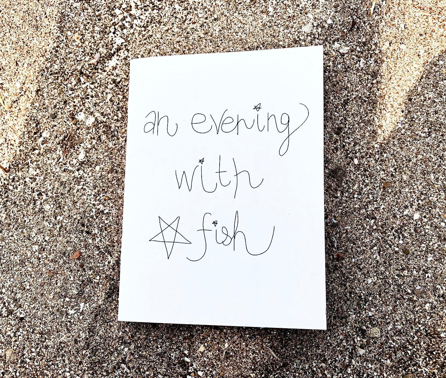 "An Evening With Star Fish" zine cover — handwritten title with small star drawing on white paper, resting on beach sand